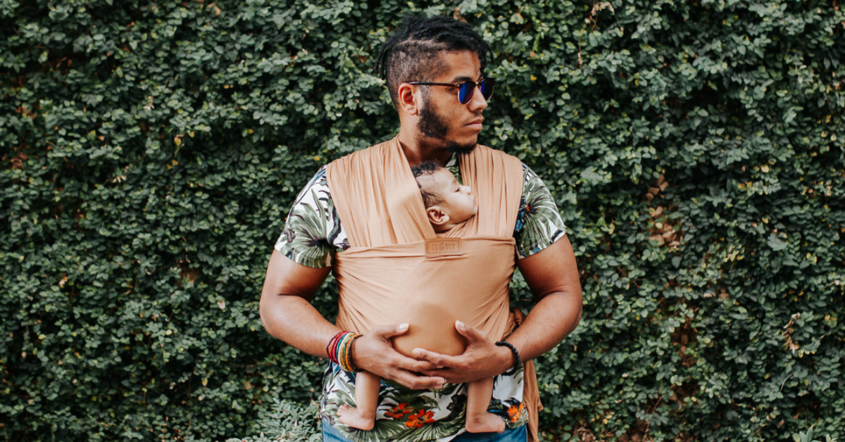 A man carries a baby in a tan wrap. He wears a floral shirt and looks off to his right. The baby is sleeping.