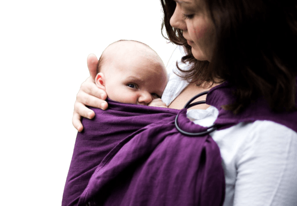 A baby in a purple ring sling looks at the camera while their parent looks down at them. 