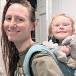 Sarah Zinn smiles at the camera. Her toddler is on her back in a baby carrier also smiling broadly.