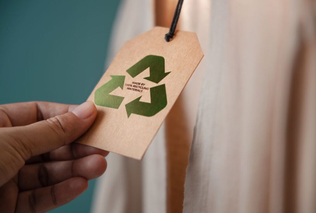 A brown hangtag with the message "100% recycled" hangs from a cloth to represent packaging for baby slings