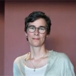 Tina Hoffman stands in front of a rose-colored wall and looks directly at the camera. She wears round glasses and has short, dark hair.