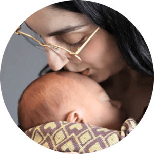 A mother holds her baby in a carrier and kisses the top of their head, demonstrating that baby should be worn high on the chest and close enough to easily kiss. 