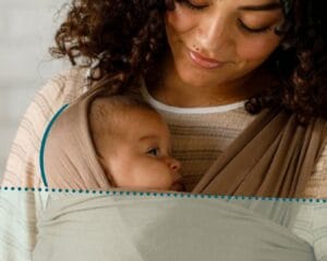 A parent looks at their baby, whose head is tucked into the wrap with the occiput lined up in a neutral position and face free from the wrap and visible to the caregiver, illustrating what safe head positioning looks like while babywearing with a stretchy wrap strap as a head support.