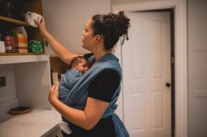 Adult puts an object on a shelf while baby sleeps in a blue wrap. 
