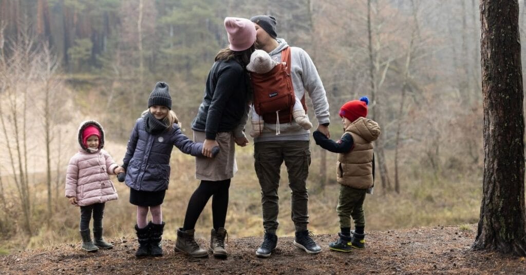 A family of six dressed in warm clothing stands in a line, with one of of the parents wearing a baby in a carrier that meets the ASTM standards for baby slings and carriers.